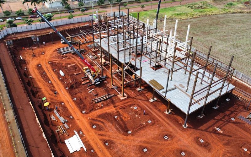 Vista aérea de canteiro: montagem de estrutura metálica com grua, laje em execução e sapatas abertas